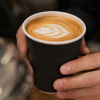 Close-up van een zwarte disposable papieren beker in gebruik, vastgehouden met beide handen en gevuld met een warme koffiedrank met melkschuim op een café-achtergrond.