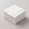 Een overzichtsfoto van een strakke, vierkante stapel witte papieren servetten op een grijze betonlook ondergrond, waarbij de ingeperste vouwlijnen in het midden van de bovenste servet een geometrisch patroon vormen.