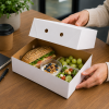 Paraatdoos 900 gram bruin/wit met losse deksel in gebruik als premium lunchverpakking. De kartonnen cateringdoos bevat een vers belegd meergranenbroodje, yoghurt met granola, druiven en een frisse salade. De verpakking wordt geopend aan een bureau- of ver