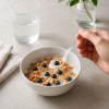 Transparante kunststof lepel in gebruik boven een kom met cornflakes, melk en blauwe bessen, vastgehouden door een hand in een heldere minimalistische ontbijtscène.