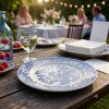 Een sfeervolle outdoor lifestyle-afbeelding van een gedekte houten tafel in een tuin. Op de voorgrond staat een leeg blauw-wit klassiek gedecoreerd bord, met daarachter een glas water en een stapel vierkante, gladde witte servetten op een ander bord. Op d