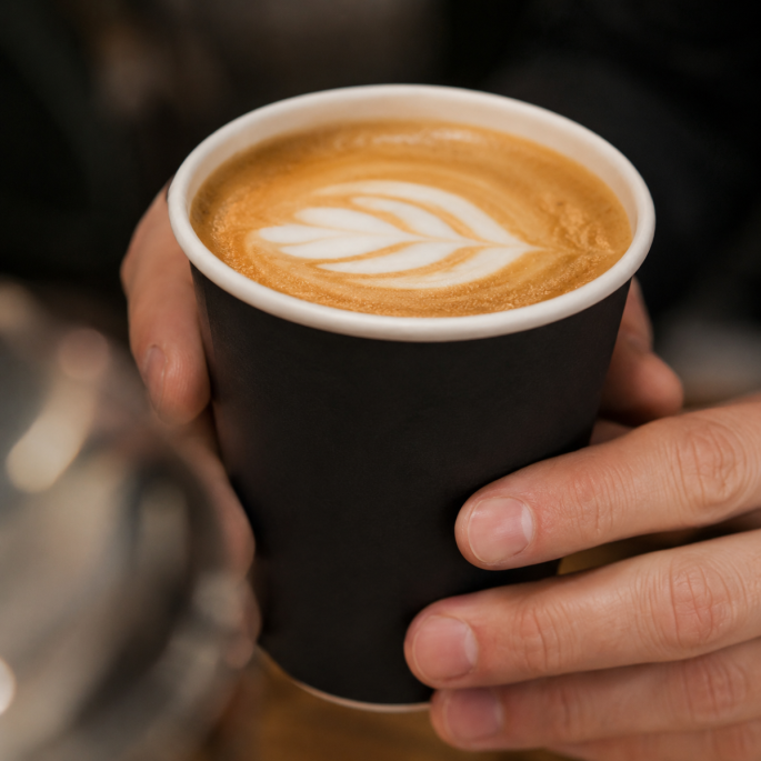 Close-up van een zwarte disposable papieren beker in gebruik, vastgehouden met beide handen en gevuld met een warme koffiedrank met melkschuim op een café-achtergrond.