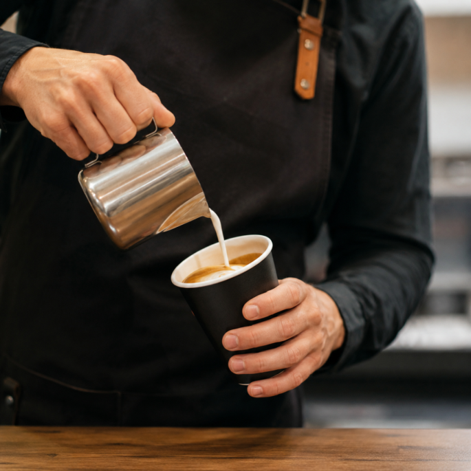 Zwarte disposable papieren beker in gebruik, vastgehouden door een barista terwijl warme melk in de beker wordt geschonken tijdens koffiebereiding.