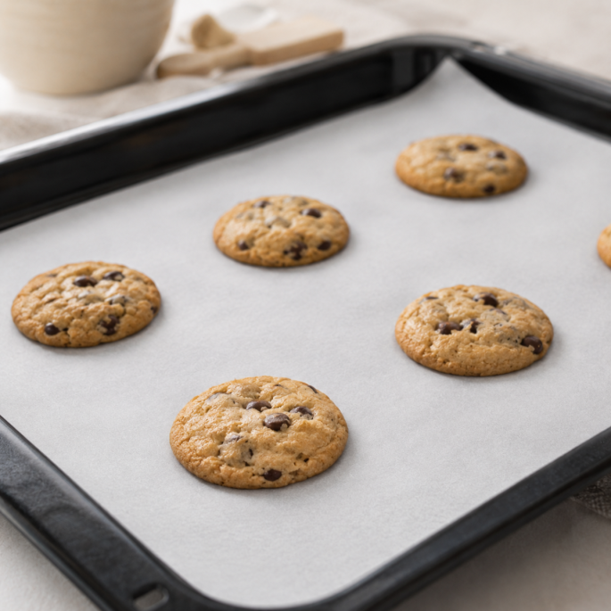 Wit gesiliconeerd bakpapier in gebruik op een zwarte bakplaat met versgebakken chocoladekoekjes, waarbij de anti-aanbakwerking en praktische toepassing van het bakpapier tijdens het bakken duidelijk zichtbaar zijn.