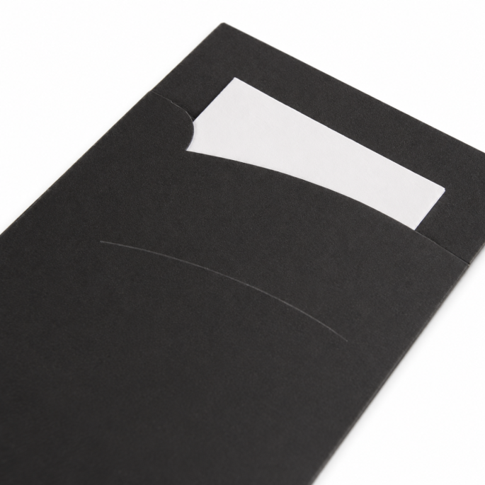 Schuin ingezoomde detailfoto van een zwarte papieren bestekhoes met matte afwerking, waarbij de gebogen insteekopening, fijne papierstructuur en het deels zichtbare witte servet duidelijk zichtbaar zijn tegen een witte achtergrond.